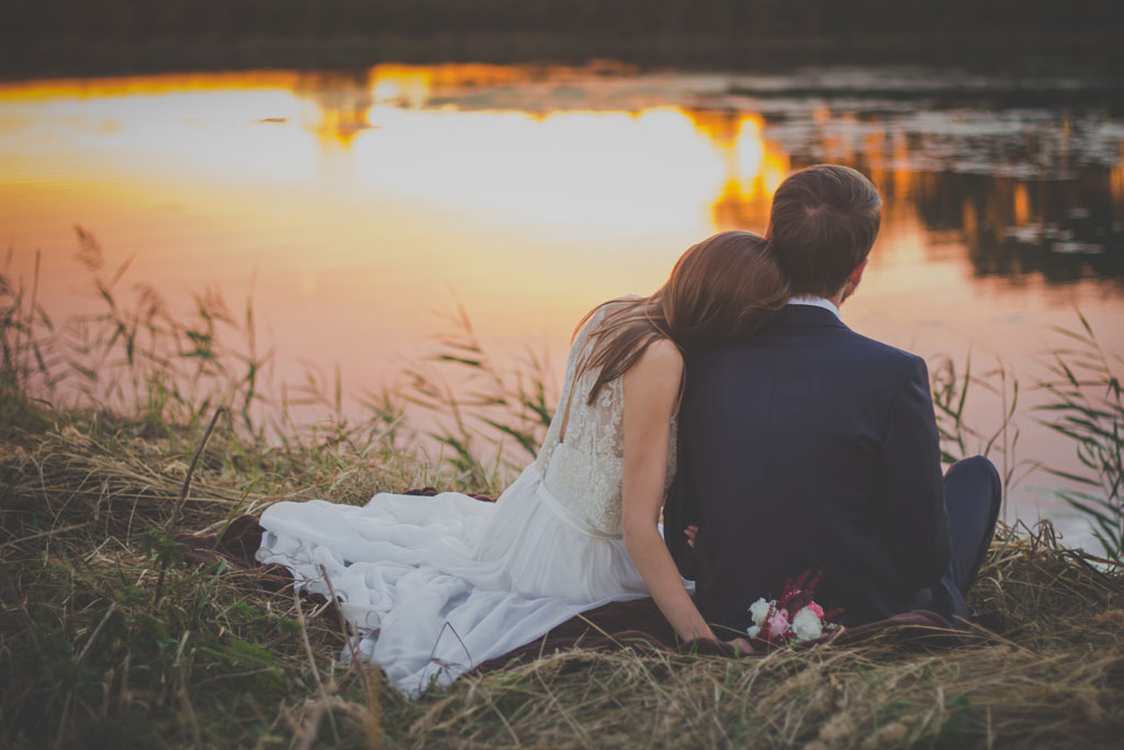 Photo of newlyweds sitting on bank of pond at sunset